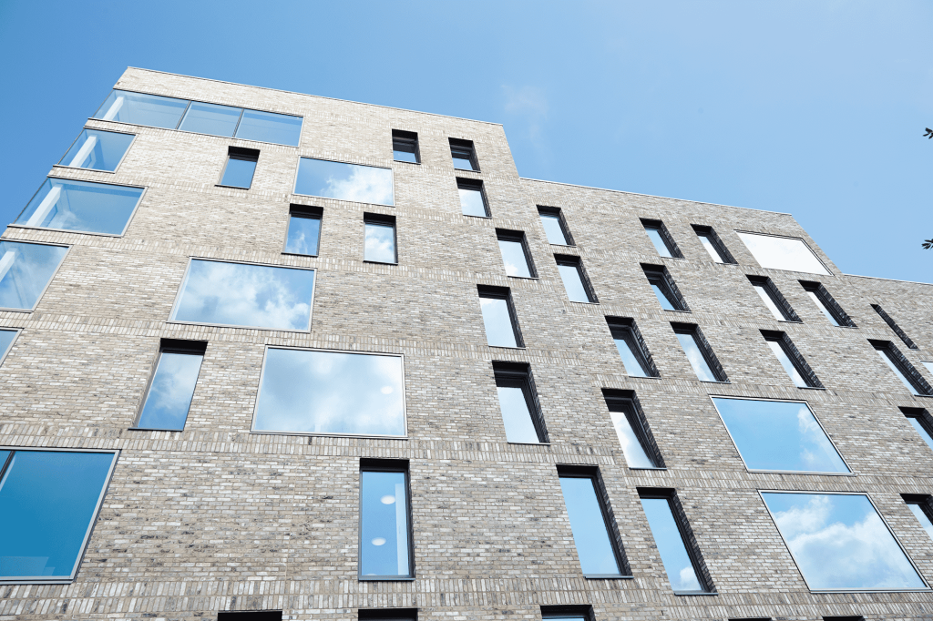 photo looking up at Norwich University of the Arts All Saints Green building shows grey brick building with several stories of windows reflecting the blue clear sky in the background