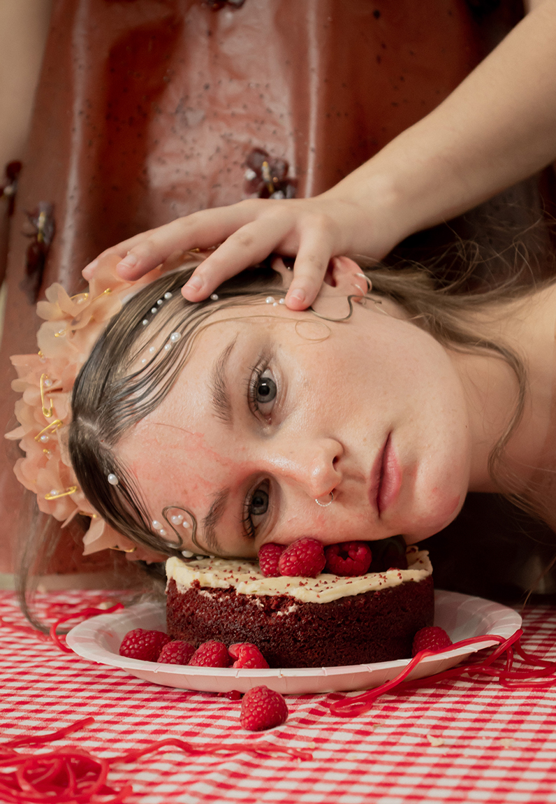 Model wears a floral pink tiara fixed together with gold safety pins, and rests their head on a slice of red velvet cake