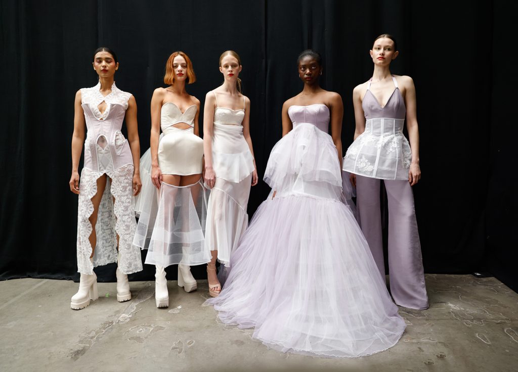 Student work by Michael Rixon for LaceyAyles. 5 models stand in a row in front of a black curtain, wearing outfits of white and lilac lace with layers, cutouts, and padding to exaggerate shape