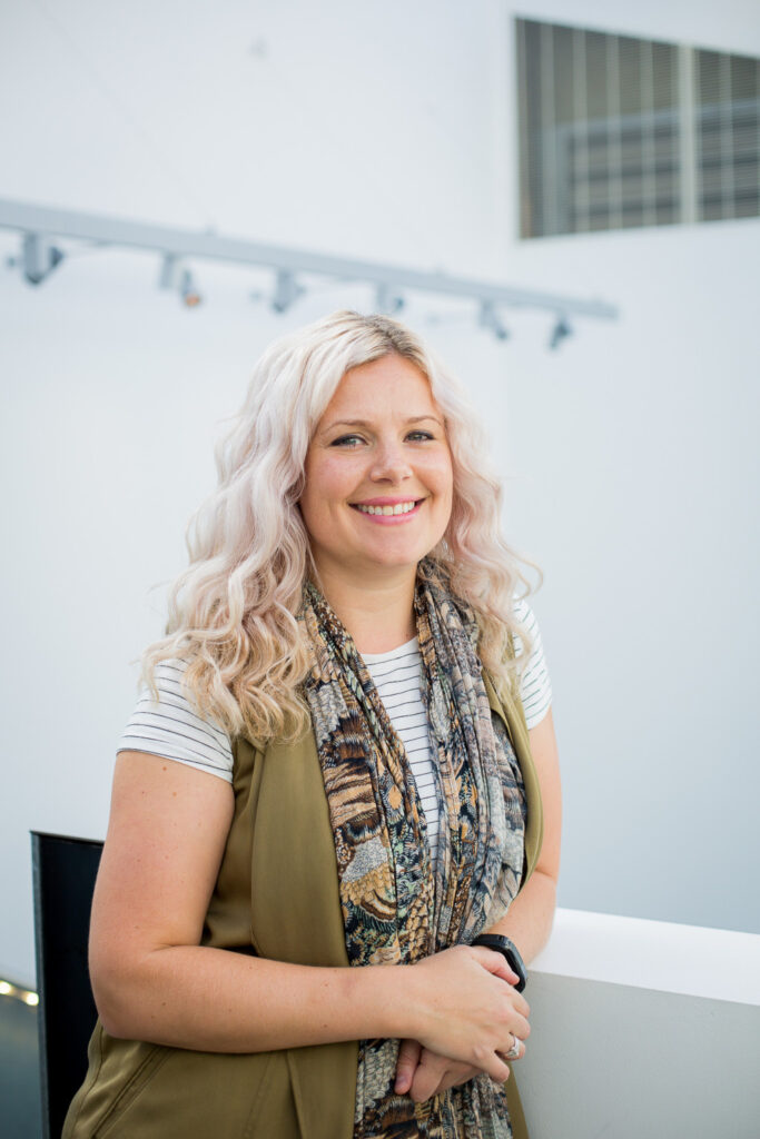 portrait photo of lecturer amy muddle standing with hands crossed and leaning on short white wall and smiling at camera with long light blonde hair and complex patterned scarf and a green sleeveless jacket