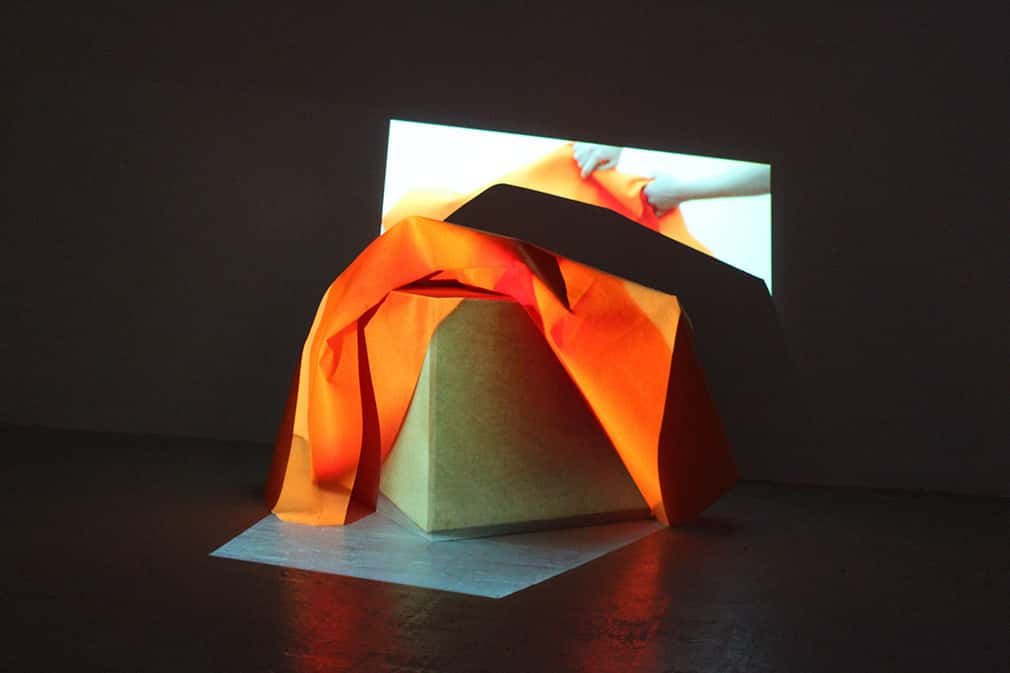 photo of exhibition piece shows lit cube with orange fabric draped over the top and a projection showing hands manipulating the same cloth being played over it