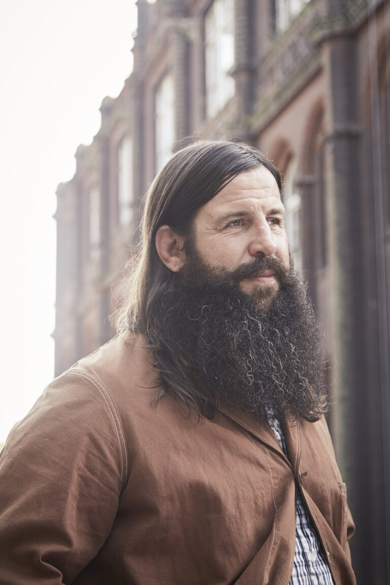 portrait photo of senior lecturer Desmond Brett standing and looking away from camera with long brown hair and a long beard wearing a light brown jacket