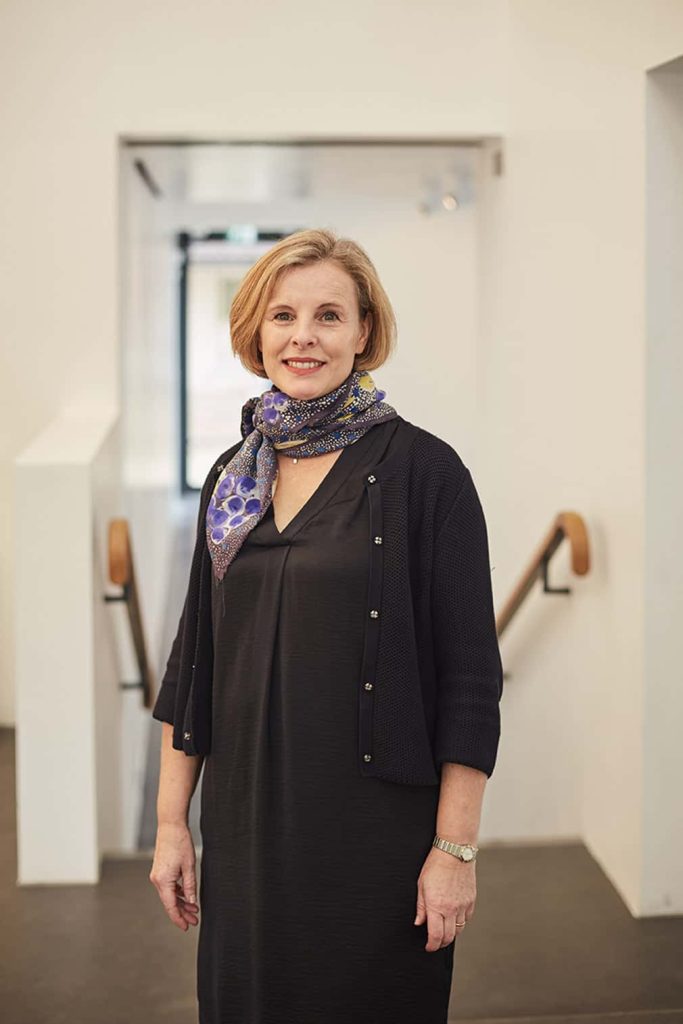 photo of doctor caroline fisher standing with arms by side and smiling at camera with medium blonde hair and a floral patterned scarf and a long black dress