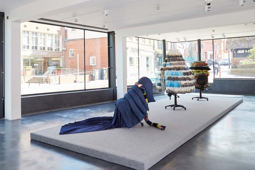 photo of exhibition at the East Gallery at Norwich University of the Arts shows a large room with dark hazy floor and tall glass windows in background and a short but wide plinth with office chairs covered in varying fabric manipulated to create folds and layers and shapes