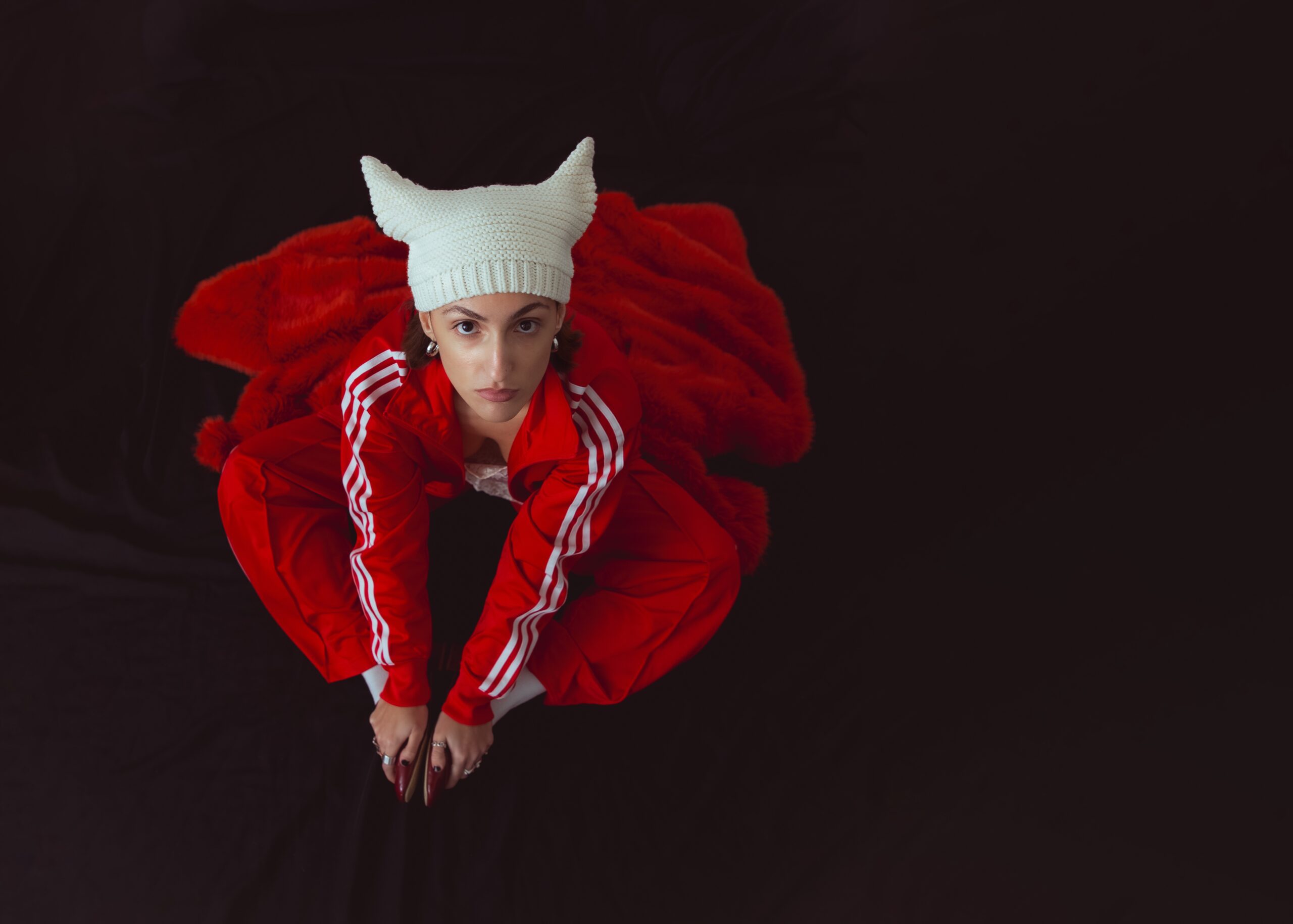 A person in a red tracksuit and white horned knit hat, embodying fashion communication marketing degree vibes, sits on a red blanket against a dark background, looking up at the camera with a serious expression.