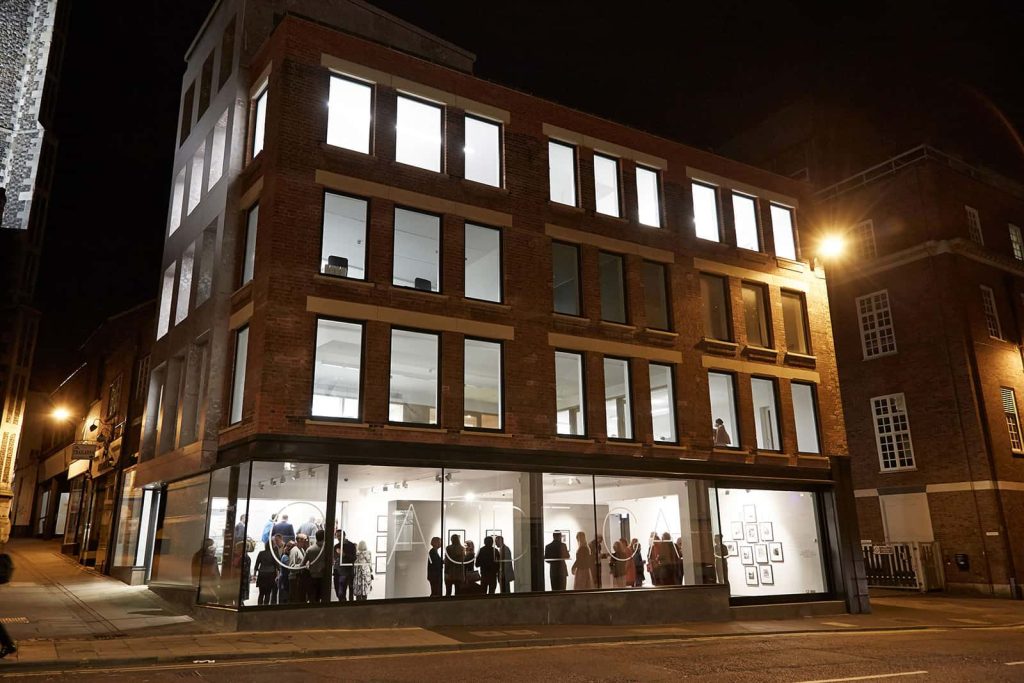 exterior photo taken at night of The East Gallery at Norwich University of the Arts shows Cavendish House with several brightly lit rooms and a gathering of people in the ground floor of the building at an exhibit