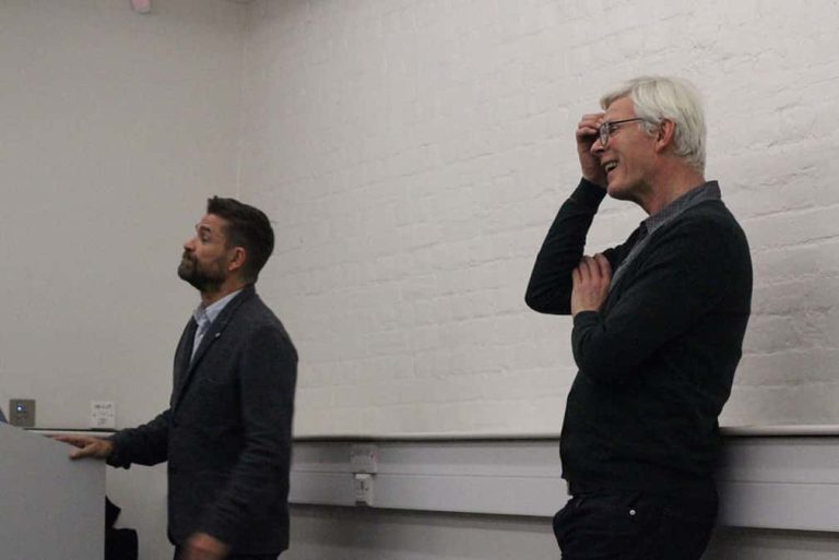 Photo of two visiting lecturers standing in front of a white wall, one is an older man rubbing his forehead with short white hair and a black jumper and the other is a middle aged man with short black hair and a beard and wearing a dark blue suit jacket