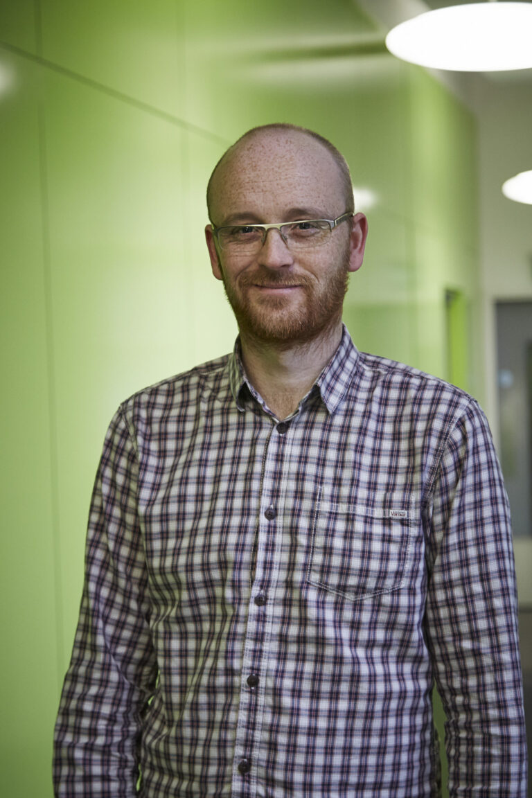 portrait photo of lecturer Jon Dunleavy standing with arms by side and smiling at camera with short red hair and a plaid white and red shirt