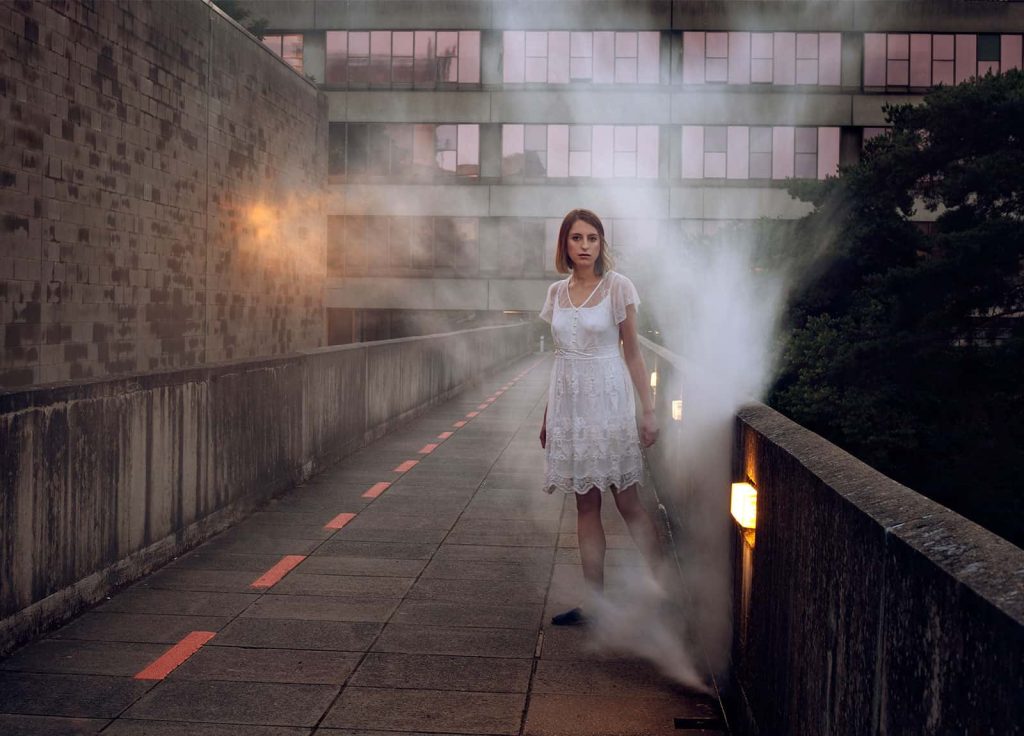 Image of a model wearing a white dress standing in a urban landscape with smoke around her