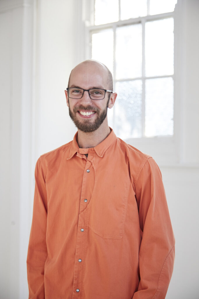 portrait photo of lecturer Matthew Benington standing with arms at side and smiling at camera with short brown hair with black square framed glasses and wearing an orange long sleeved loose shirt