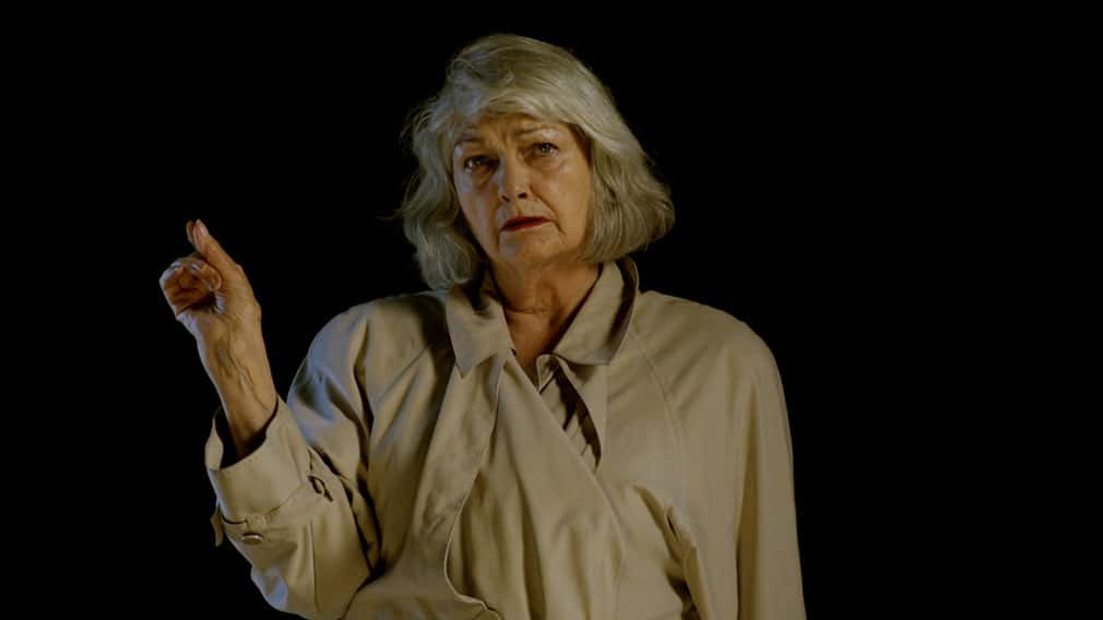 A woman with medium length grey hair and a beige raincoat pointing a finger