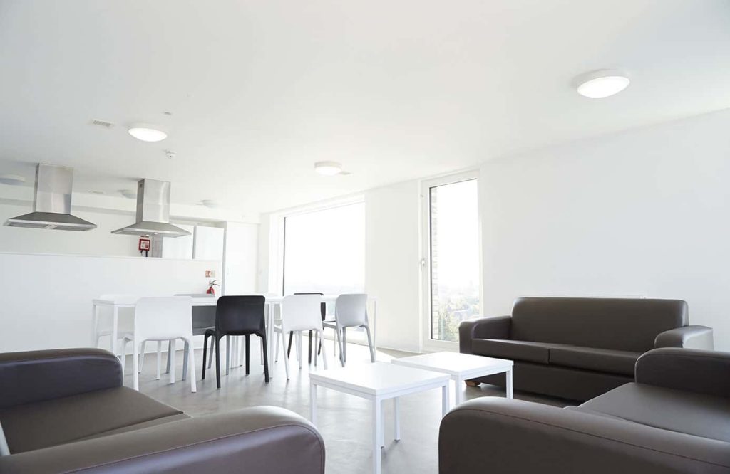 photo of wide white room with tall and wide floor length windows with brown leather sofas in foreground, a white table and chairs in the midground and extractor fans for the kitchen in the background