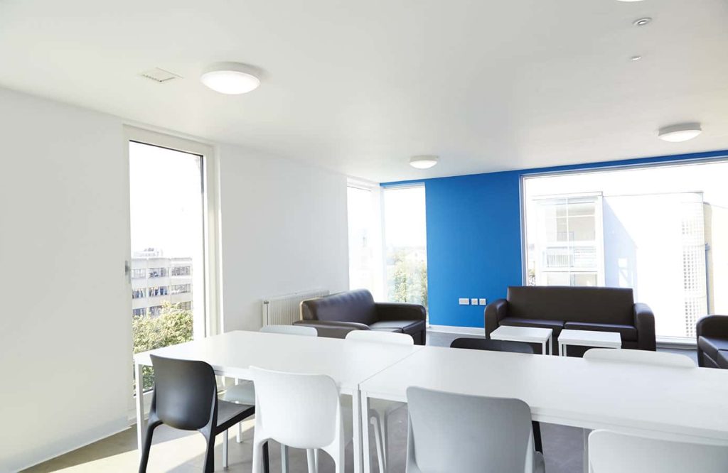 photo of wide white room with blue feature wall and wide floor length windows with white table and chairs in foreground and brown leather sofas in background