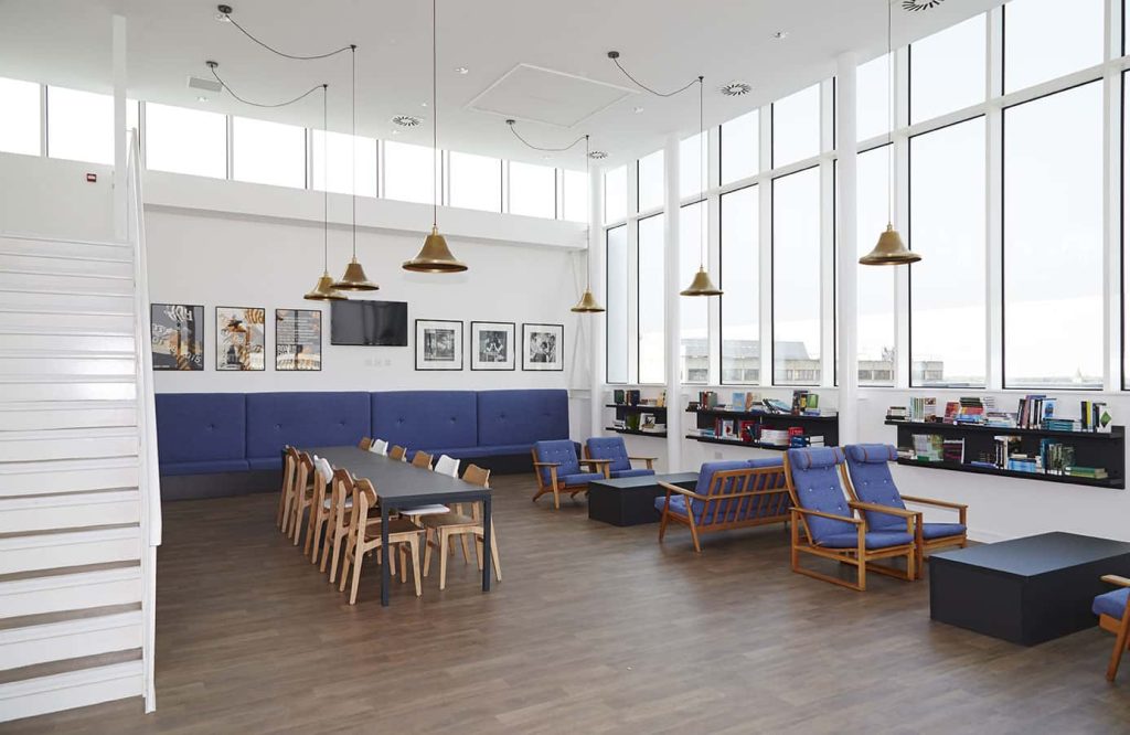 interior photo of Norwich University of the Arts building All Saints Green building shows high ceiling room with white walls and wooden floor with chairs, tables and shelves with books and high windows letting in bright light with a view of Norwich.