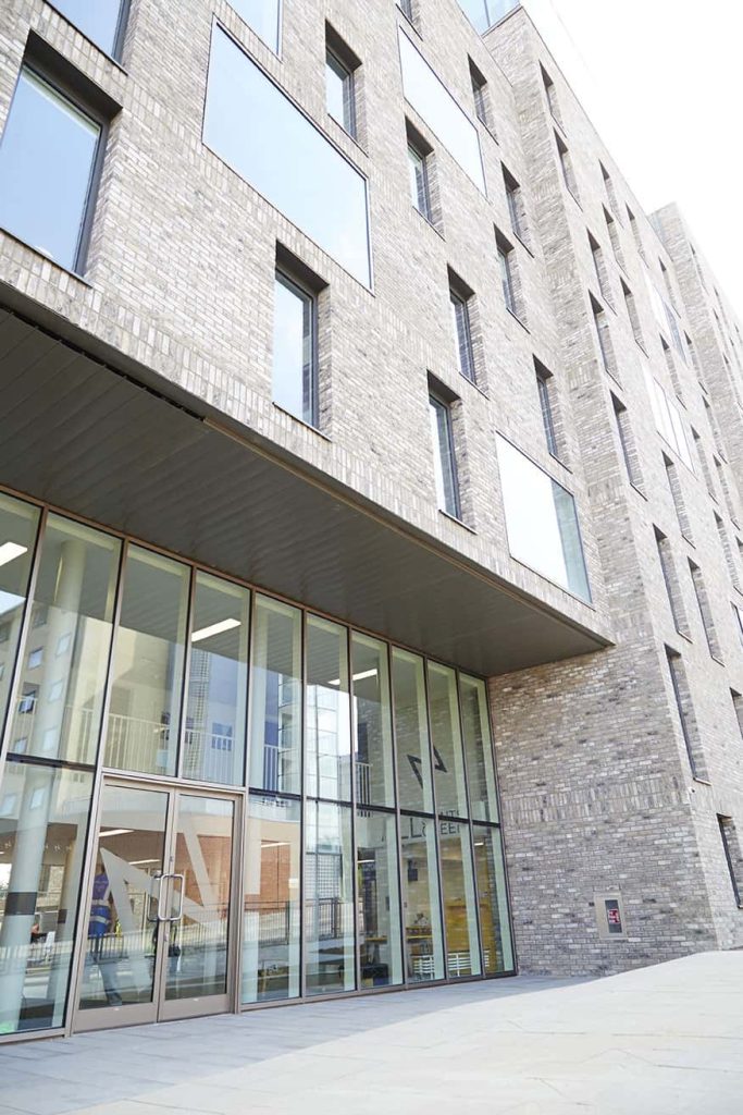 photo shows Norwich University of the Arts All Saints Green building with grey brickwork and many windows reflecting the bright blue sky with ground floor glass entrance