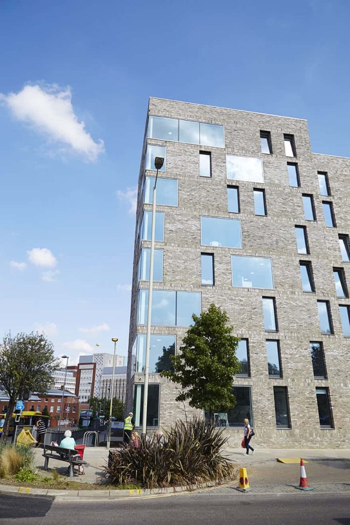 photo looking up at Norwich University of the Arts All Saints Green building shows grey brick building with several stories of windows reflecting the blue clear sky in the background