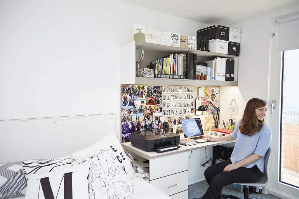 photo shows a white bedroom with a tall window on the right hand side and a student sitting at a desk with a board of several images and paperwork and a shelf of storage items and a bed with cushions