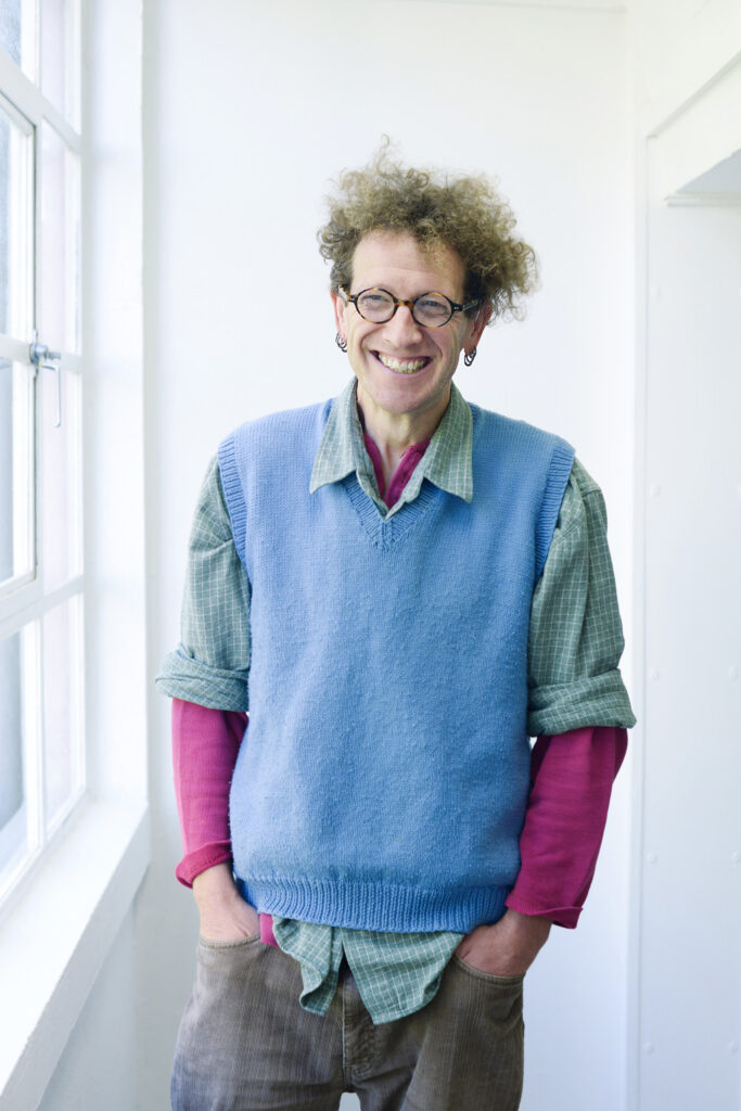 portrait photo of lecturer Nigel Bousfield standing with hands in pockets and smiling at camera with curly blonde hair and round brown glasses with a blue sweatervest and plaid green and white rolled sleeve shirt and a long sleeve pink undershirt