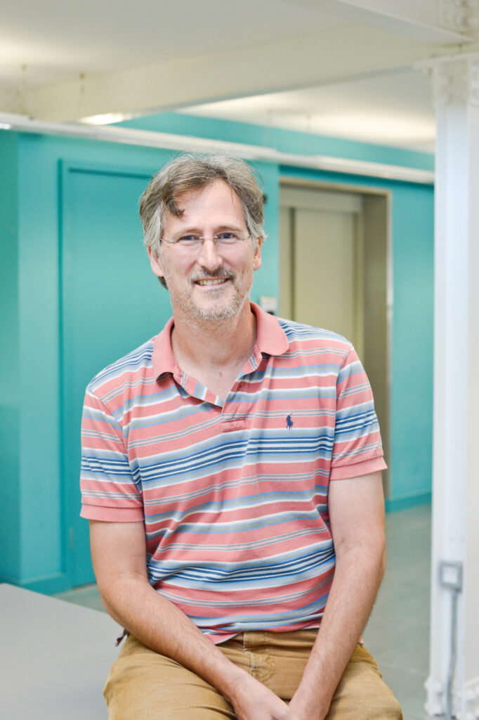 portrait photo of senior lecturer doctor nicholas maffei sitting on a desk with hands in lap and smiling at camera with short light brown hair and silver glasses and a pink and blue horizontal striped shirt