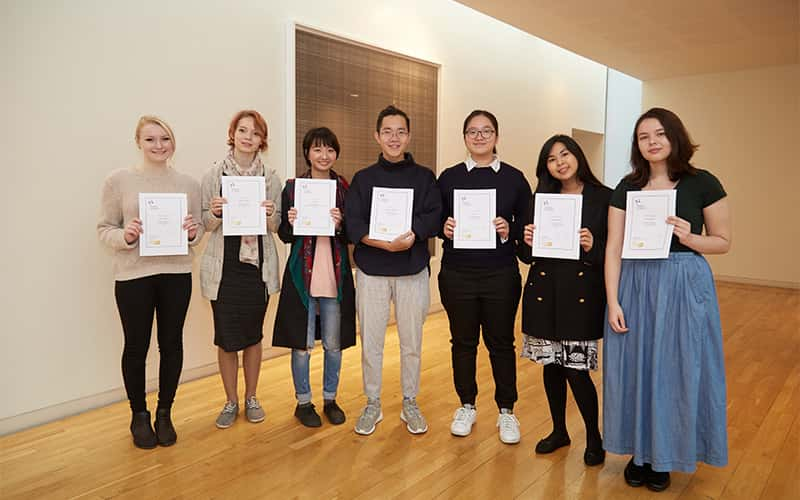 photo shows seven students standing adjacent all holding award certificates to camera and smiling in a wooden floored white room