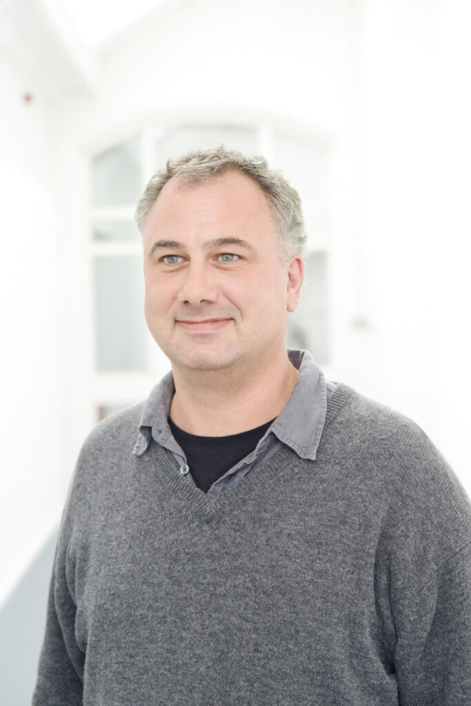 portrait photo of year zero co-ordinator Shaun Camp smiling and looking away from camera with short grey hair and a grey long sleeved jumper