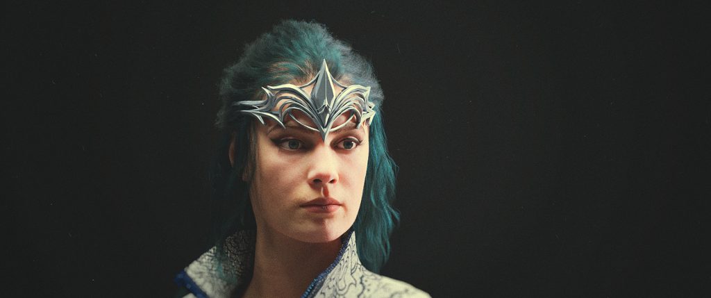 A woman with blue hair and an intricate silver crown-like headpiece faces forward, slightly looking to the right. Her elaborate outfit, complete with a high collar and detailed designs, could easily be a project in an animation visual effects degree course. The dark background makes her stand out prominently.