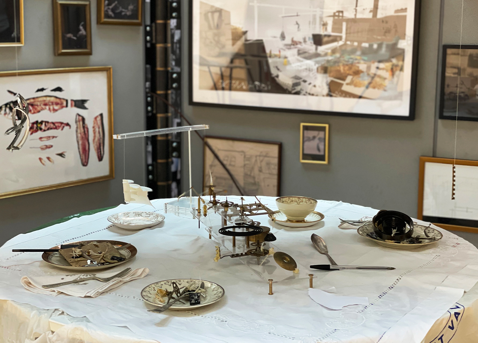 Student work by Theo Galvin shows architectural model on a set dinner table surrounded by gold framed artwork