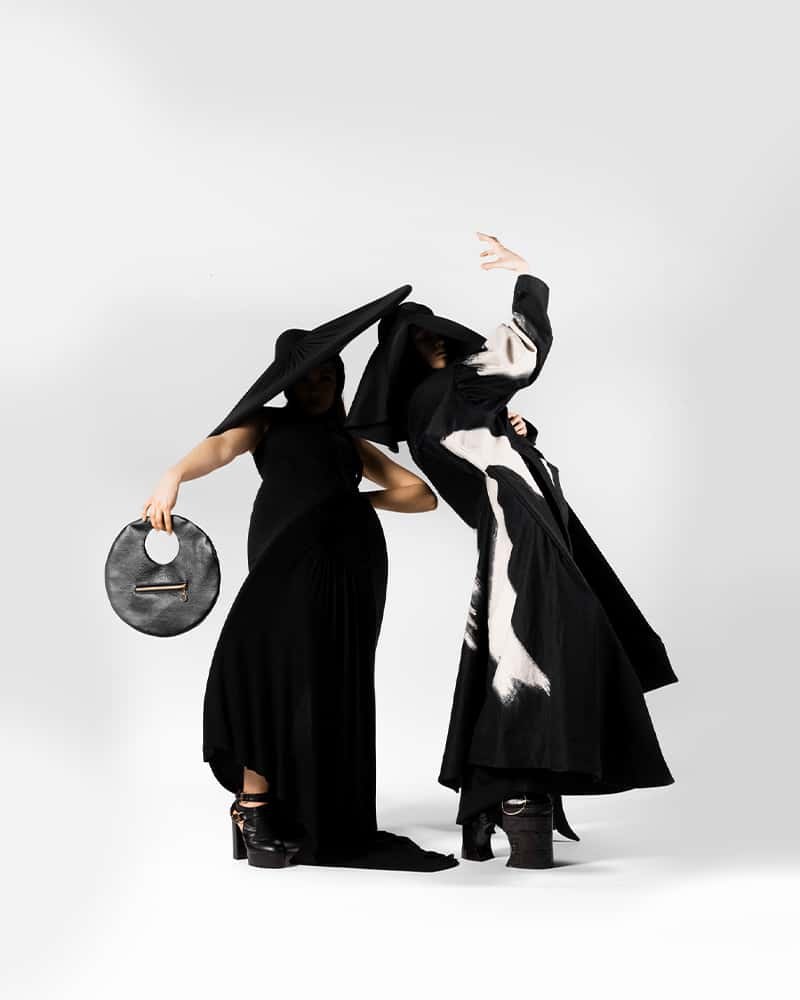 Two figures strike a pose in long monochrome dresses, with their faces obscured by the shadows from their extremely wide-brimmed hats. Together, their silhouette is almost a circle on the white background