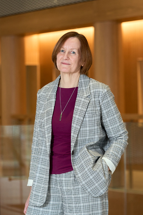 Hilary Carlisle stands wearing a grey check suit with burgundy top, in a room with wood panelling.