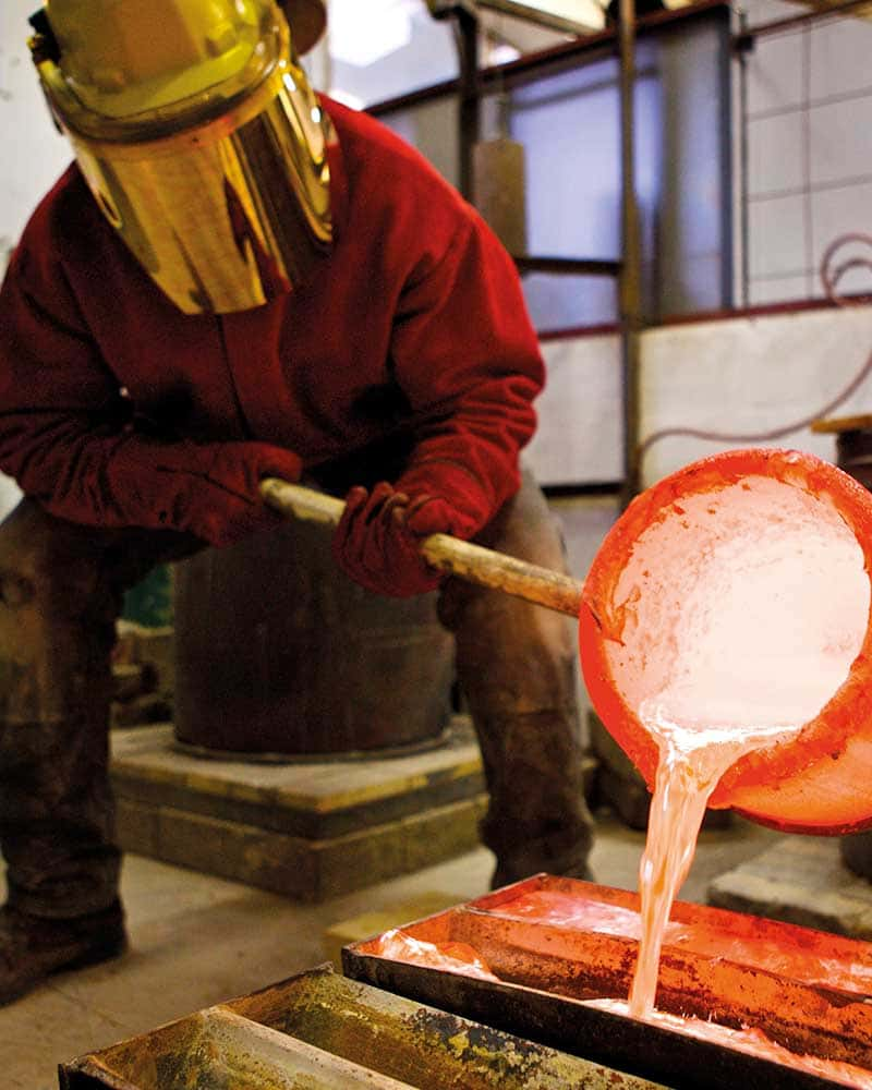 Image of a man wearing a gold protective mask is pouring molten metal into a cast