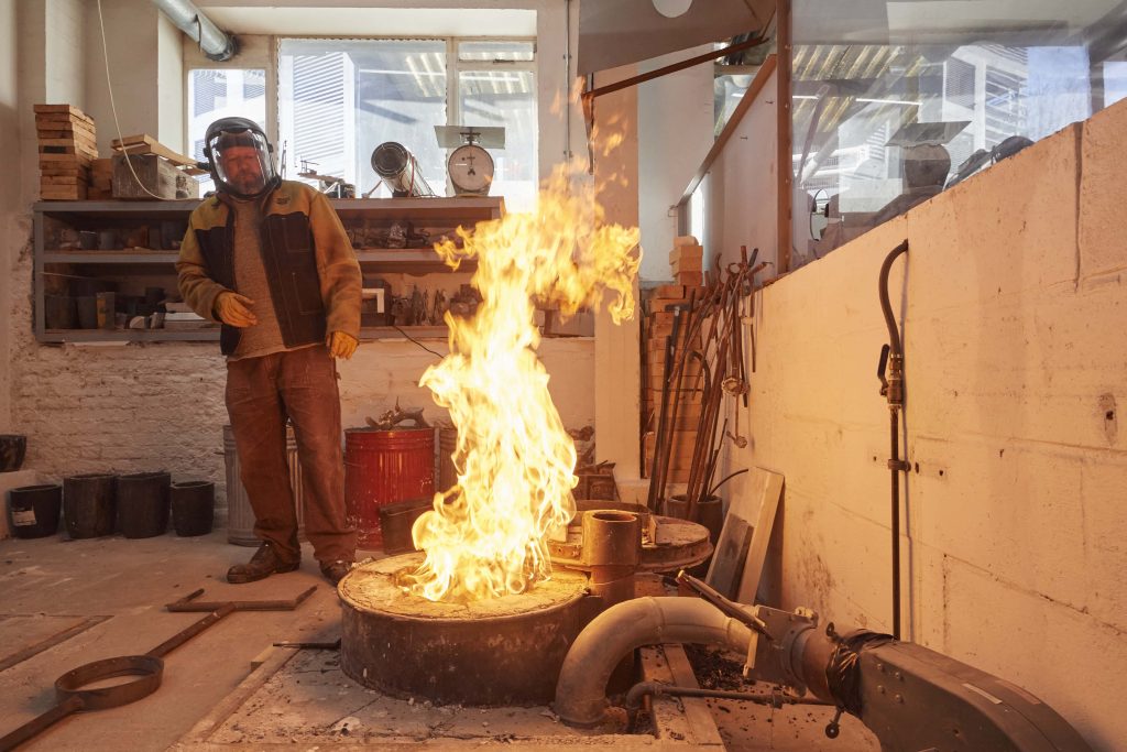 A technician doing a demonstration of 'the pour' in the foundry in the 3d workshop watches a large (controlled) flame while pouring molten metal to make a sculpture