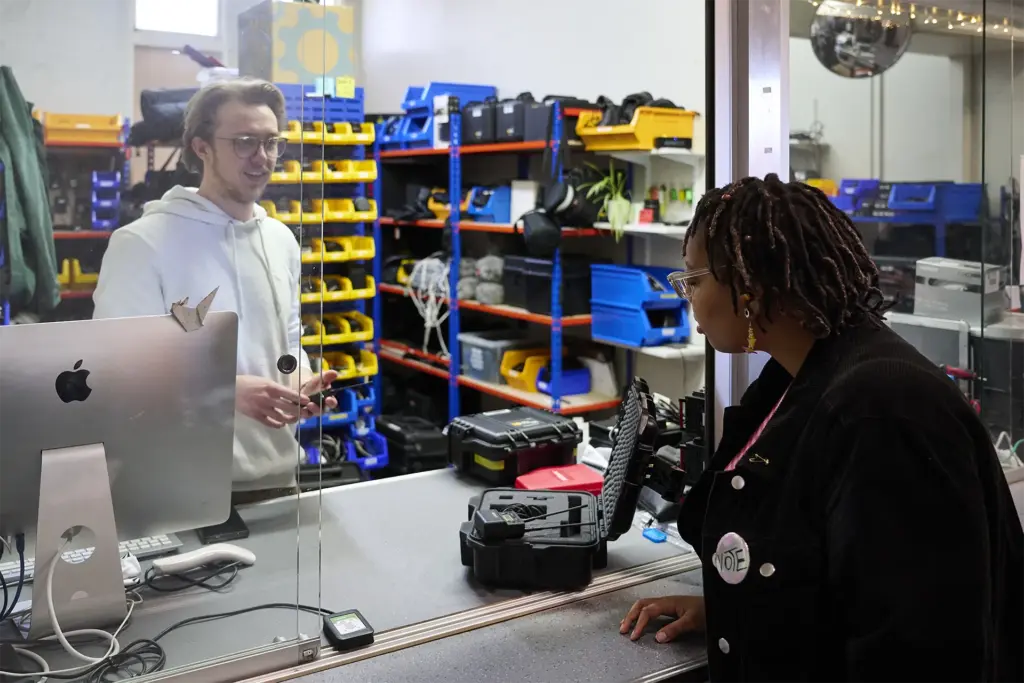 A person in a white hoodie stands behind a counter with computer equipment, talking to another person wearing glasses and a "VOTE" button in front of the counter. Shelves with bins and tools are in the background.