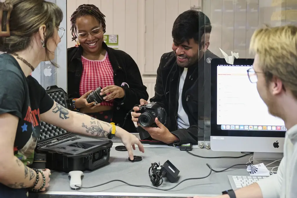 Four people gather around a table with camera equipment and a computer, smiling and examining a camera together in a bright room. One person is opening a case while others look on and interact.