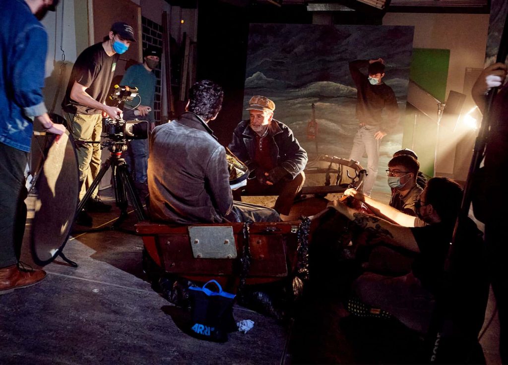 An old sailor sits in a rowing boat with a short grey beard. The boat is being hand rocked by students who crouch out of shot, in front of a large painted backdrop of stormy skies and seas. The sailors short peaked hat, jacket, and fingerless gloves have been drybrushed with white paint around the edges and details which makes him look slightly like he is a painting, and a beam of harsh warm light obscures some details in the photo.