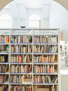 The library is a serene haven where a tall white bookshelf holds assorted books. Sunlight streams through arched windows, creating a warm atmosphere. In the background, more shelves and cozy reading spaces are partially visible, inviting endless exploration in this literary sanctuary.