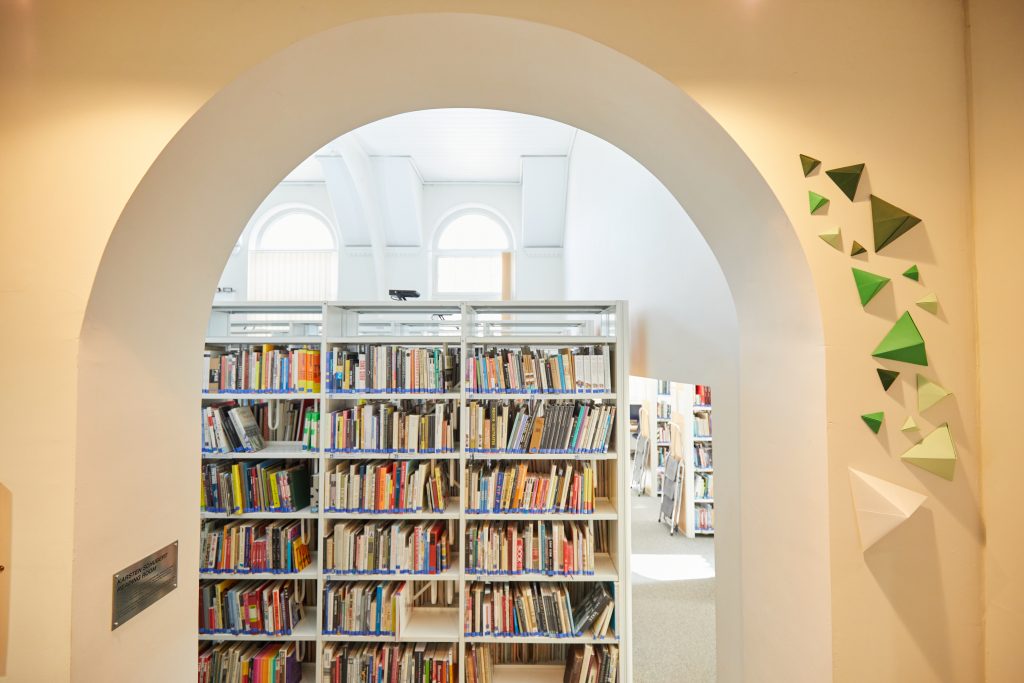 A serene library interior unfolds through a large archway, with bookshelves brimming with knowledge. The light-colored walls feature geometric decorations in green and white. Bright, natural light filters through the arched windows, illuminating this sanctuary of learning.