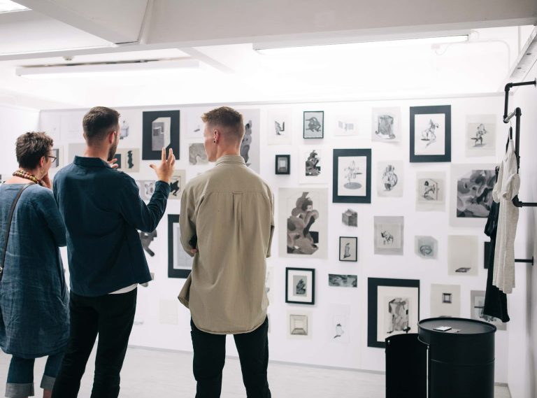 Three people looking at a wall full of framed drawings in the 2017 BA Degree Show