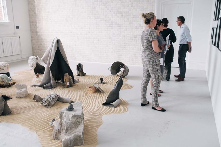 Sculptures in the middle of the room on sand with people looking at work on the walls