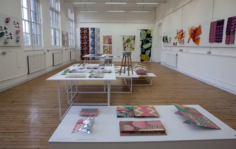 Large room with wooden floor displaying textile design work on tables and walls in 2017 BA Degree Show