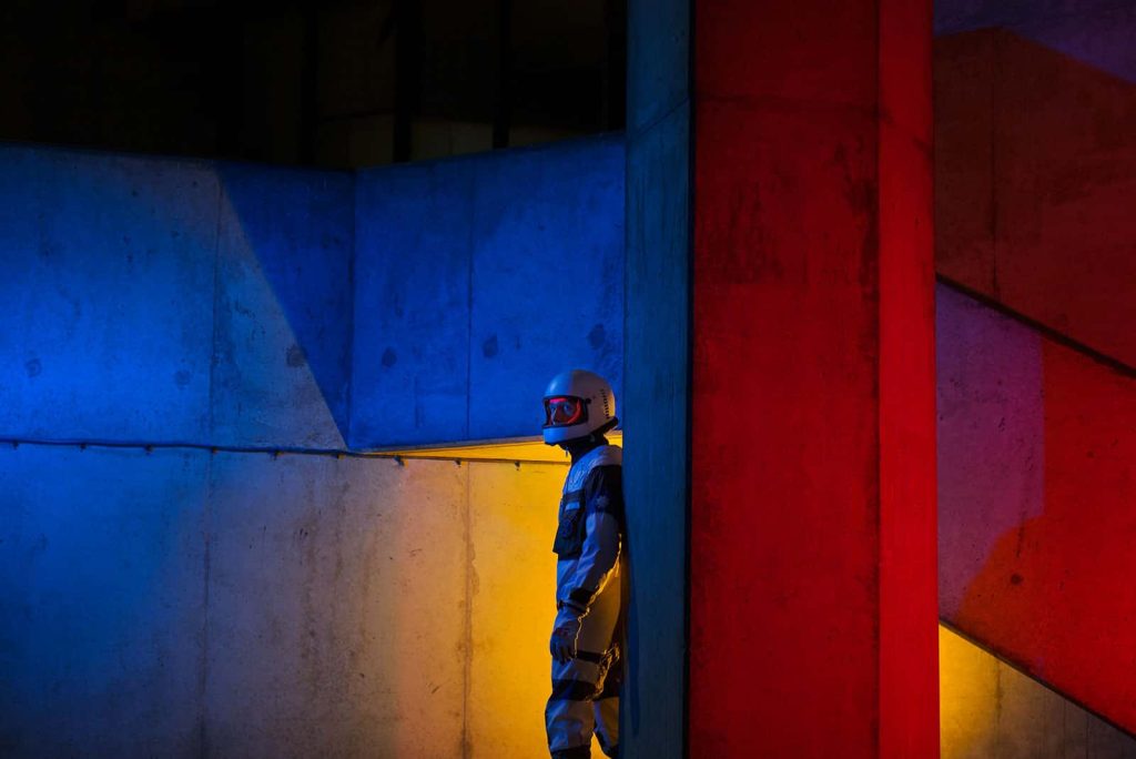 Image depicts a man in a space suit standing in a concrete scene with red, blue and yellow lights projected onto the space