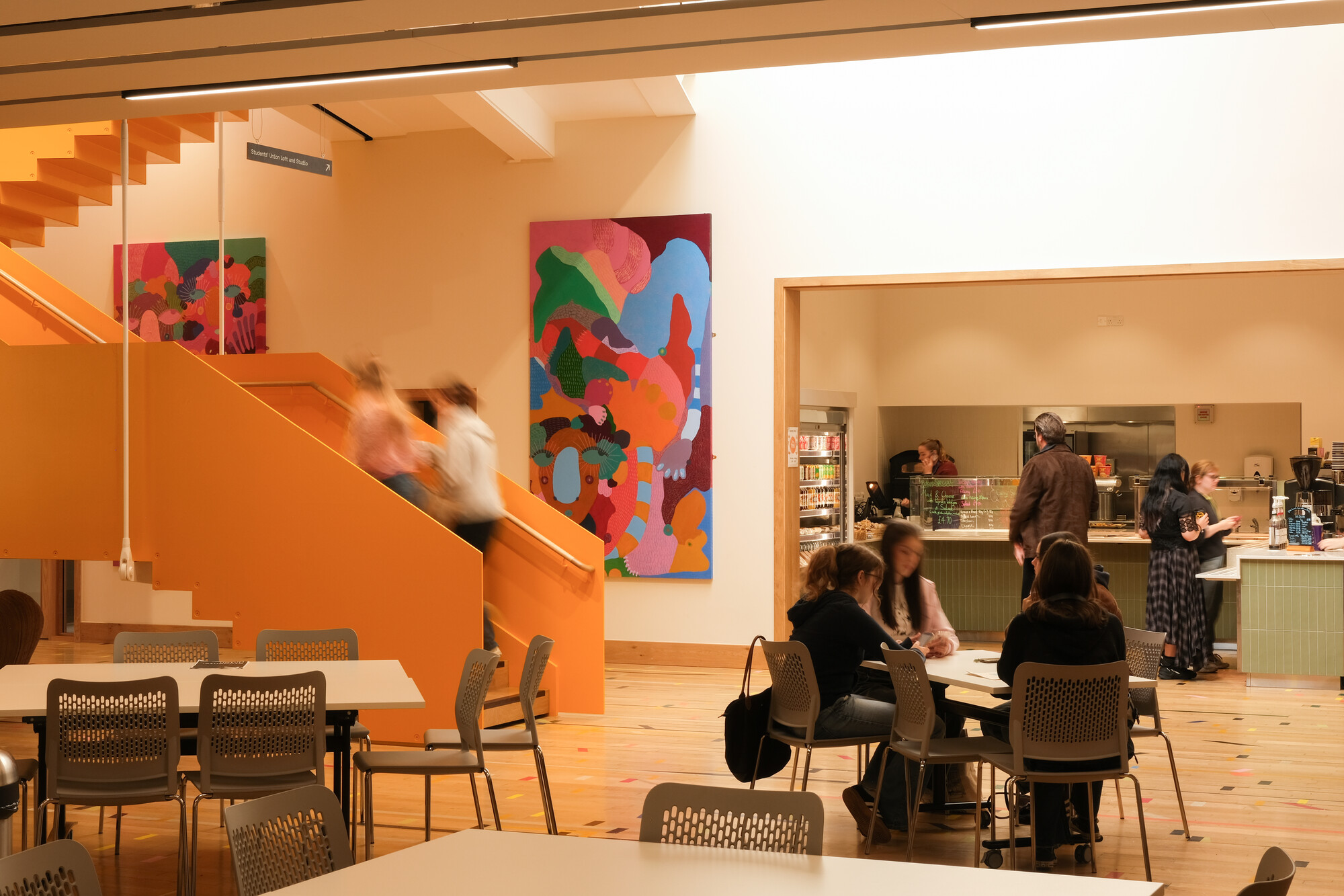 A brightly lit cafeteria with orange stairs, colorful abstract artwork on the walls, people ordering at a counter, and others sitting at tables talking or working.