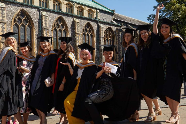 Graduates from Norwich University of the Arts in graduation gowns stand outside St Andrews Hall