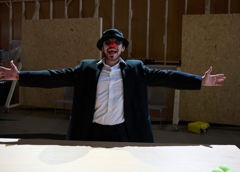 A person in a dark suit and hat, wearing face paint, stands behind a table with arms outstretched and mouth open wide. Their theatrical presence hints at an acting degree. The background features unfinished wooden panels and scattered tools, while the focused lighting creates a dramatic effect.