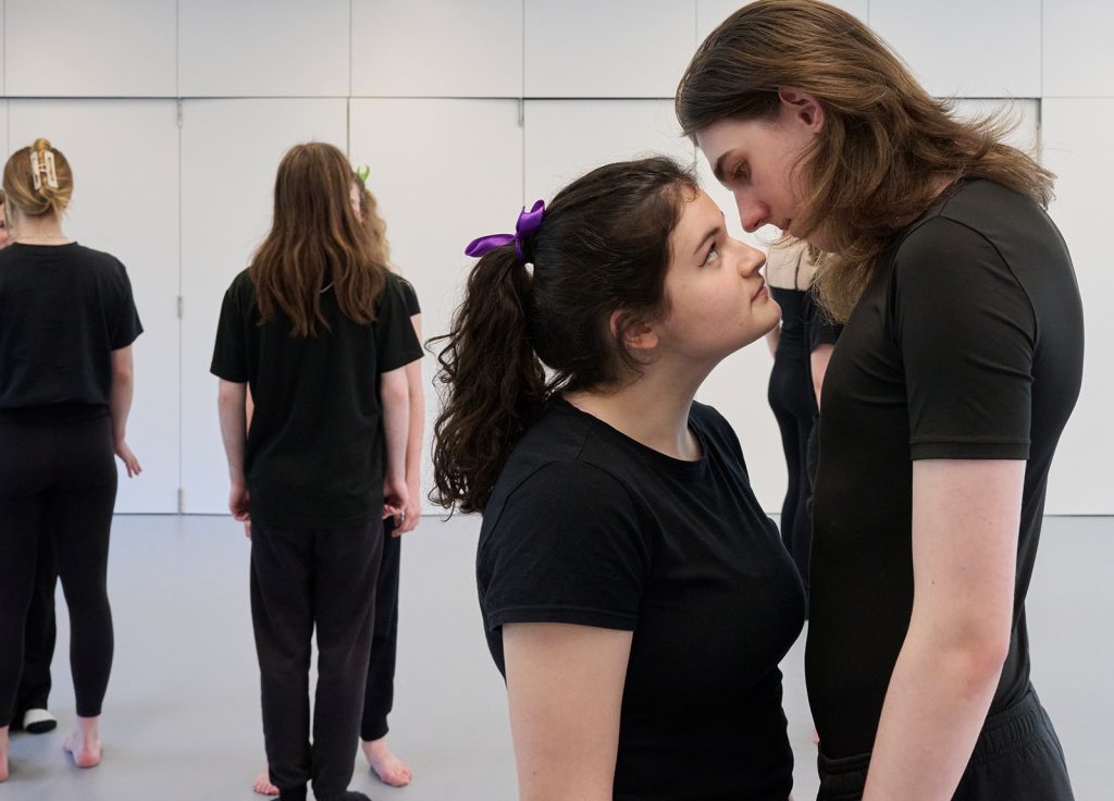 Four people in a room, all dressed in black, showcasing their skills from an acting degree. In the foreground, two individuals stare intently at each other, while in the background, the other two stand facing a white wall. The room has a minimalistic design with white walls and gray flooring.