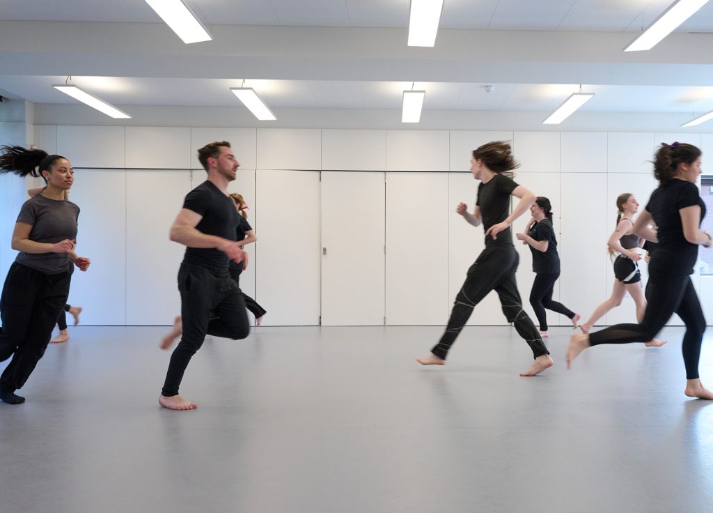 A group of people wearing casual athletic attire run barefoot in a spacious, well-lit room with white walls and ceiling lights. They appear to be participating in a movement class, likely part of an acting degree program.