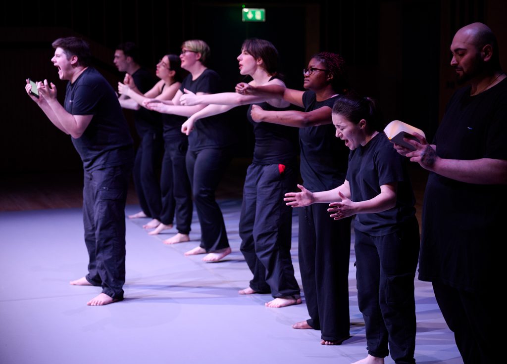 A group of barefoot individuals, wearing black, perform expressive gestures while standing in a line on stage. The green exit sign glows in the background. Each person, trained through an acting degree, is engaged in dynamic movements, creating an atmosphere of intense, synchronized performance.