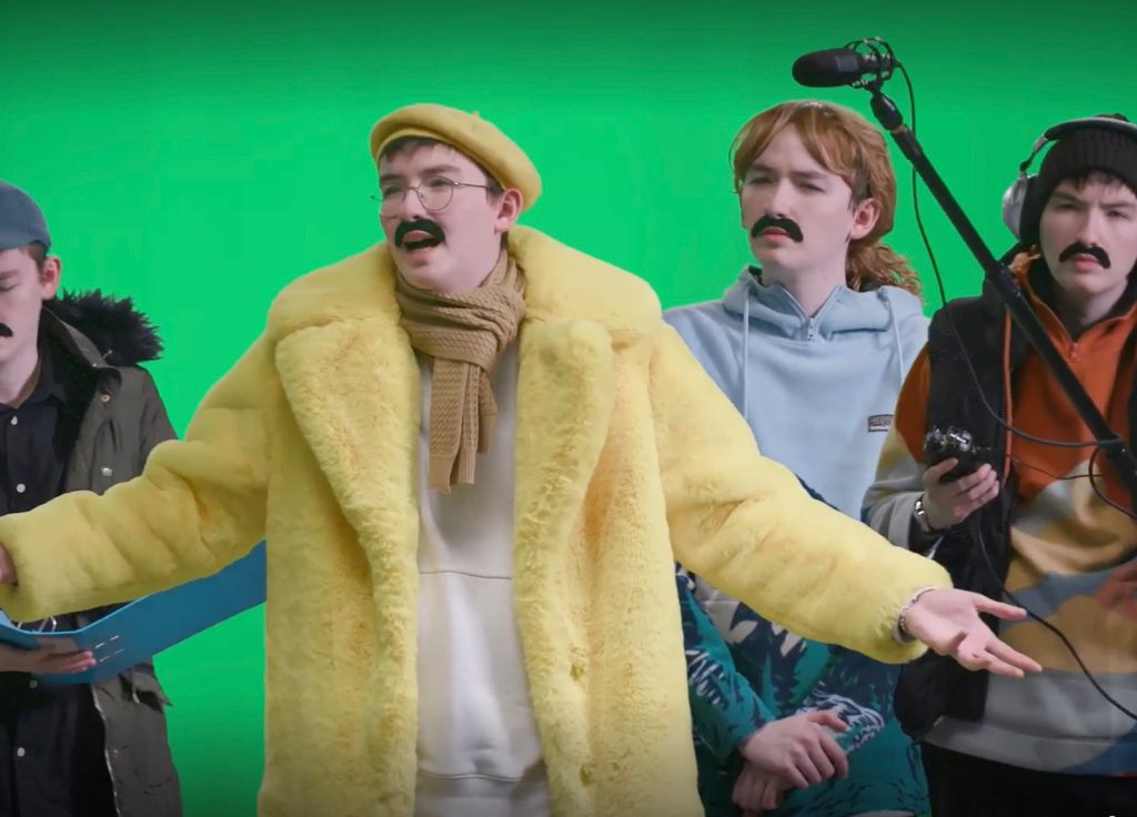 A group of four people stands in front of a green screen, all wearing large fake mustaches. The person in the center, possibly flaunting an acting degree, dons a bright yellow fur coat, yellow beret, and light brown scarf. The others are casually dressed, including one holding a boom microphone.