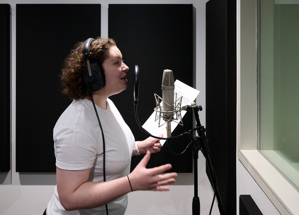 A person with curly hair, wearing a white t-shirt and headphones, stands in a recording studio speaking into a microphone. Holding a sheet of paper, they appear to be passionately engaged in their performance, showcasing the skills honed from an acting degree.