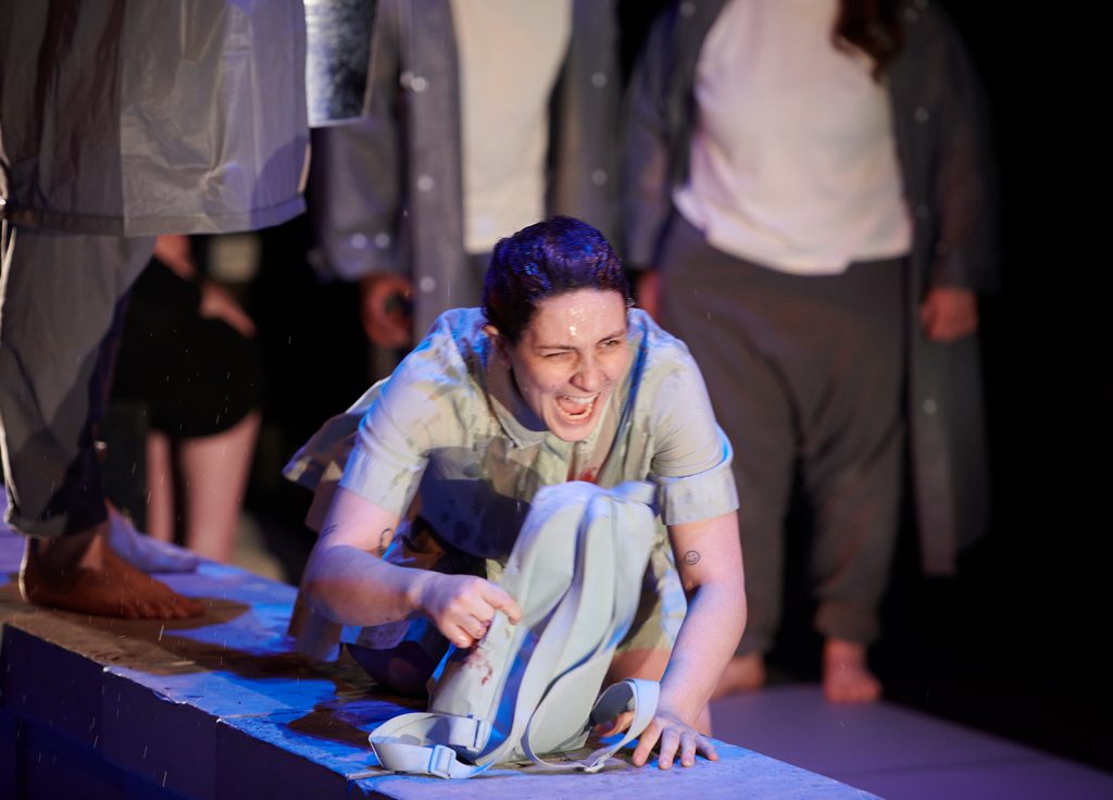 An acting student crawls on the ground carry a bag with bloody handprints on it infront of several people wearing baggy clothes and barefoot