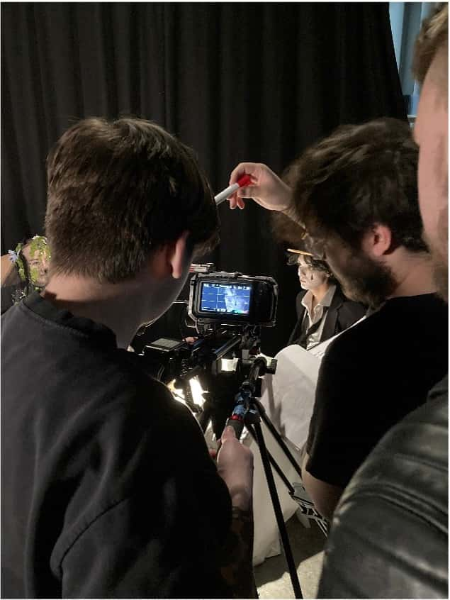 Film and acting students collaborating to product a music video. Image is over the shoulder of 3 students watching a performance through the screen of a camera on tripod